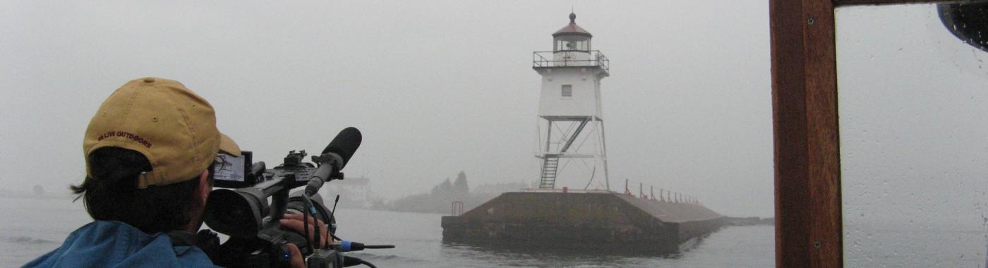 Back of a man wearing a yellow cap, holding a camera and filming a lighthouse in the foggy distance