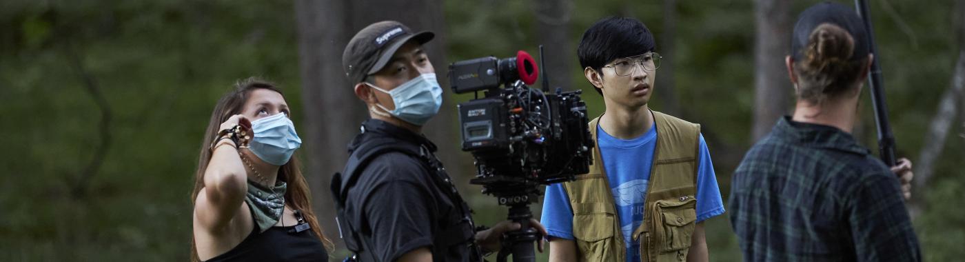 A woman and a man with a camera both wearing surgical masks. A younger Asian American man wearing a vest and the back of a man with a boom mic.